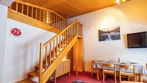 En trätrappa som leder upp till ett loft, en matplats med ett bord dukat för två personer och en väggmonterad tv.