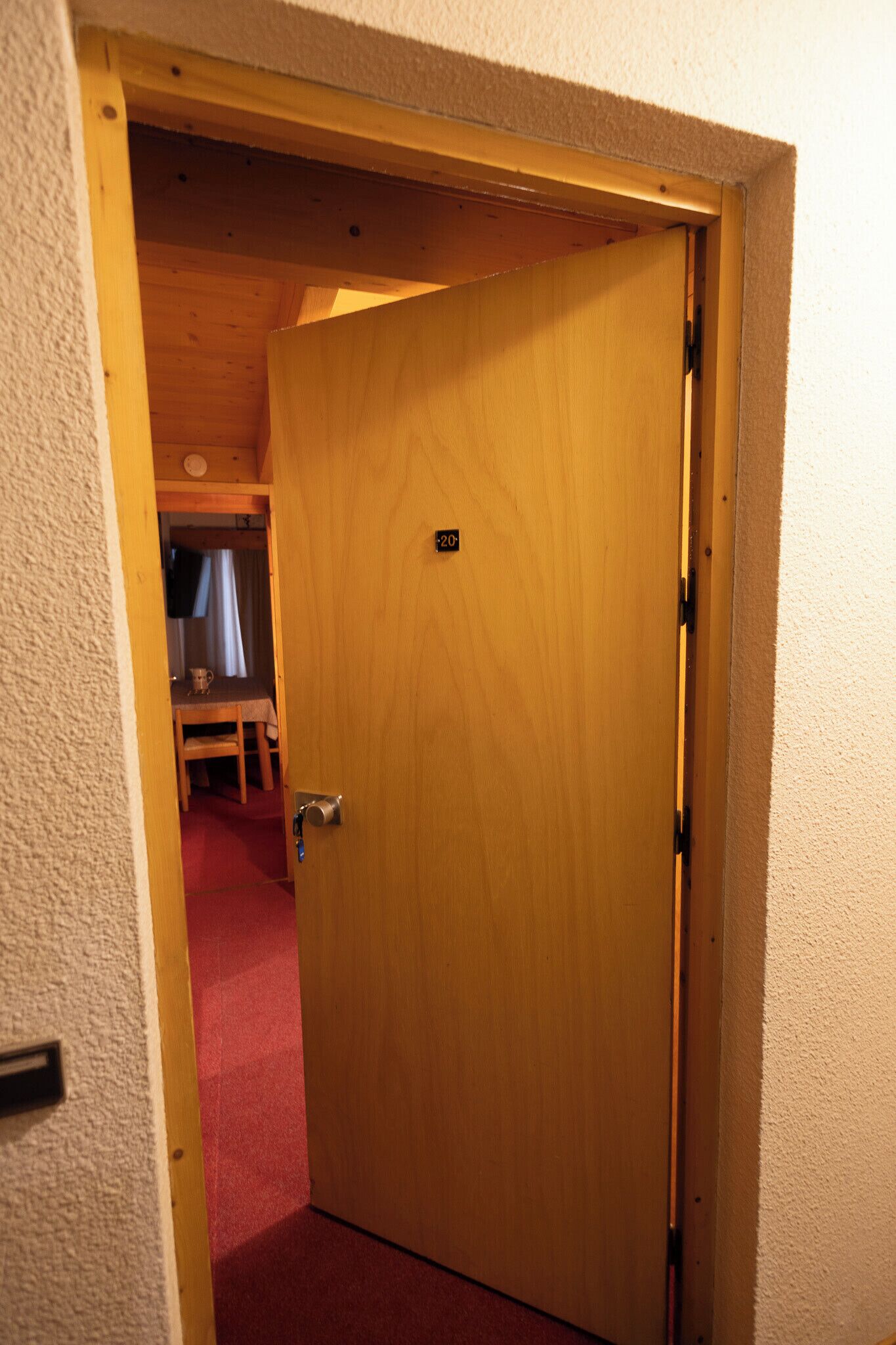 Une porte en bois, ornée d’une plaque portant un numéro, donne sur une pièce où se trouvent une moquette rouge et une table en bois, ainsi qu’une chaise.