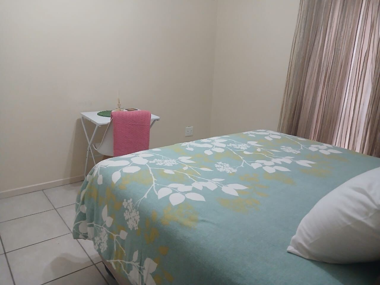 A bedroom with a bed, a small table with a pink towel, and a window with curtains.