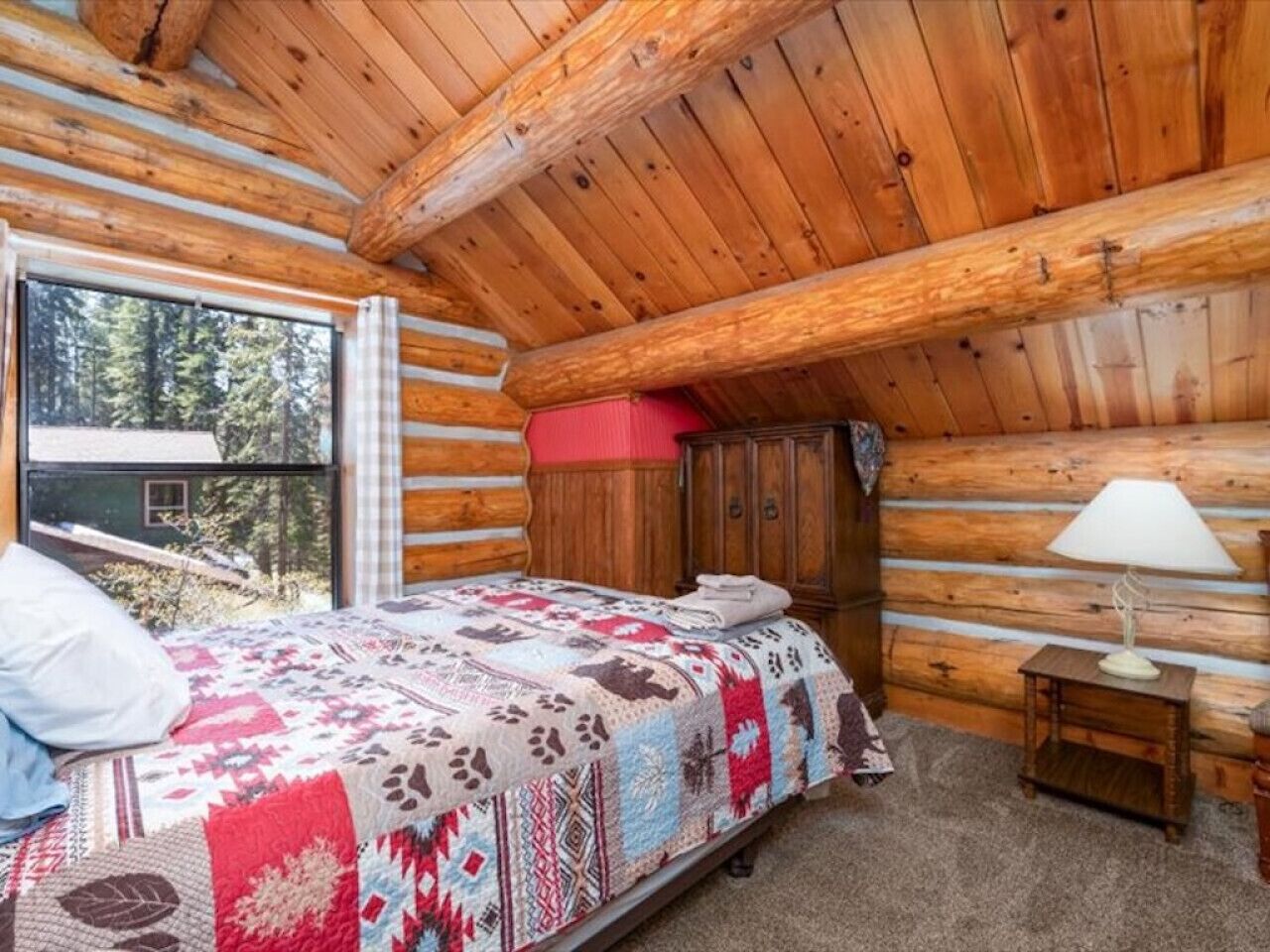Un dormitorio tipo cabaña de madera con una cama, una mesita de noche, una lámpara y una ventana con vista a árboles.