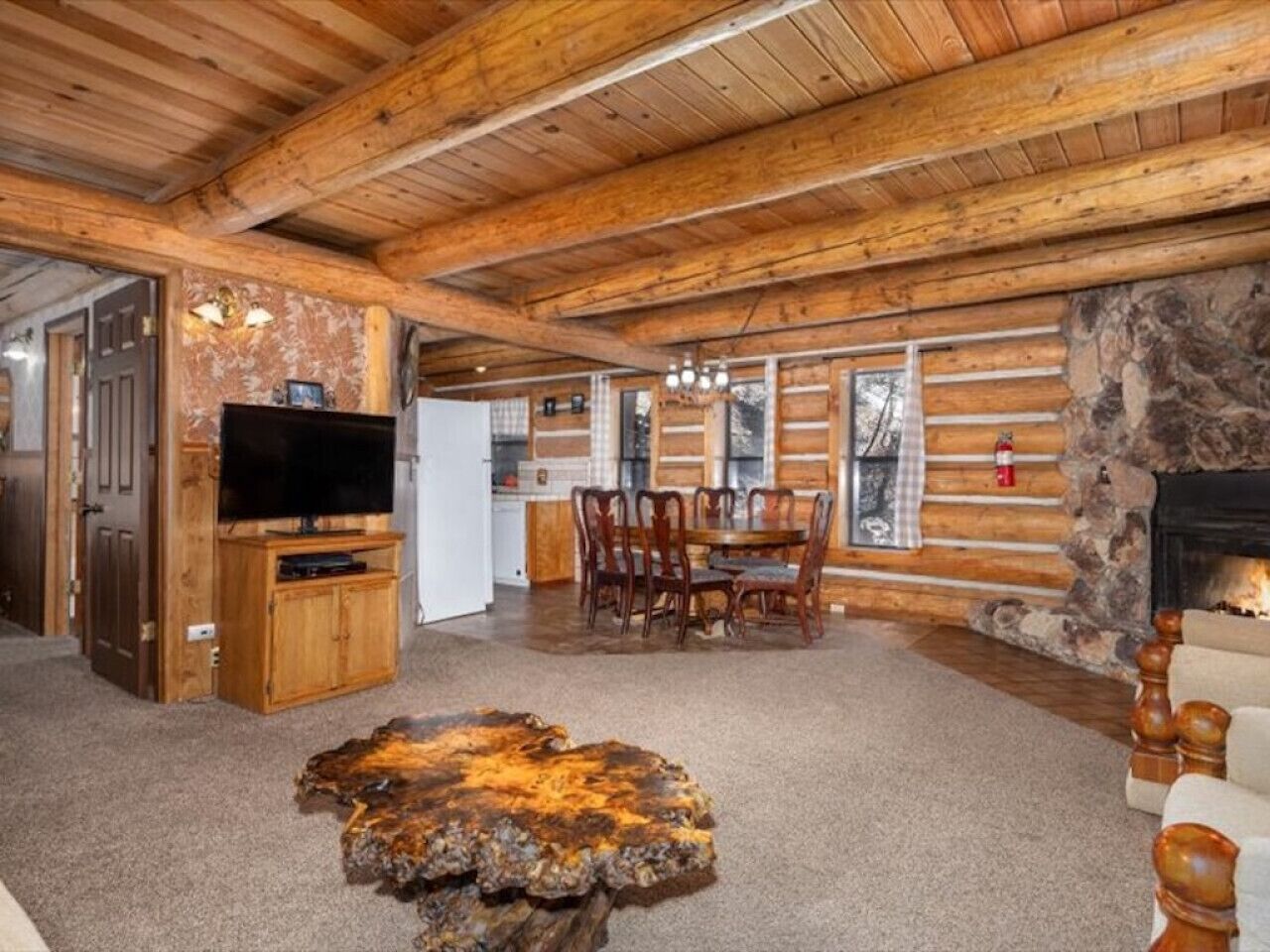 A log cabin interior with a stone fireplace, wooden dining table, and a television in the living area.