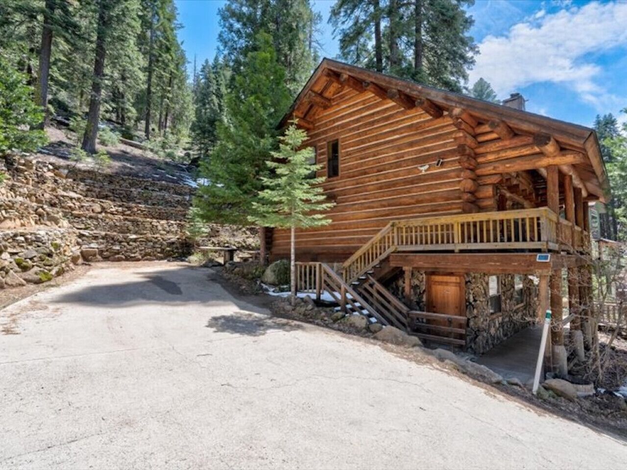 A log cabin with a porch and stairs leading to a deck, surrounded by trees and a stone wall.