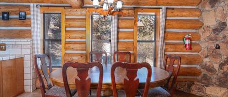A wooden cabin interior with a dining table surrounded by chairs, a chandelier, and a stone wall.