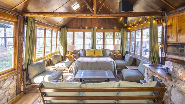 Una cabaña con interior de madera, una cama, zona de estar y ventanas amplias.