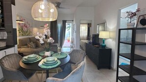 Un salon moderne avec un coin repas, comprenant une table ronde avec des sets de table verts, un luminaire suspendu en osier et une vue sur l’extérieur grâce à une grande fenêtre.
