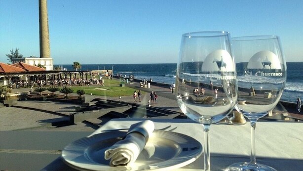 Une table avec deux verres à vin et une serviette roulée, donnant sur une zone côtière avec un phare et une promenade.