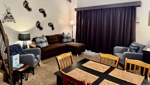 Una sala acogedora con un sofá modular, una mesa de comedor con individuales y un ventanal con cortinas oscuras.