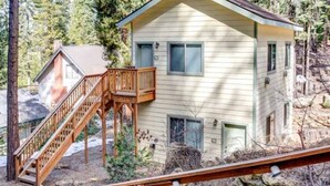 Una casa de dos pisos con una escalera de madera que conduce a una terraza, rodeada de árboles y terreno rocoso.