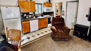 Cocina compacta con armarios de madera, microondas y refrigerador. En la sala hay un sillón marrón y una silla de madera.