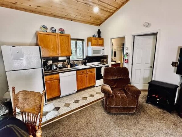A compact kitchen with wooden cabinets, a microwave, and a refrigerator. There is a brown armchair and a wooden chair in the living area.