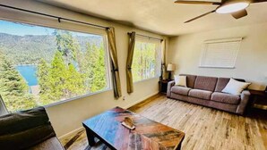 Ein Wohnzimmer mit Couch, Holztisch und Blick auf See und Wald durch große Fenster.
