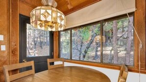 Une table à manger en bois, des chaises, un lustre et une vue sur les arbres à travers de grandes fenêtres.