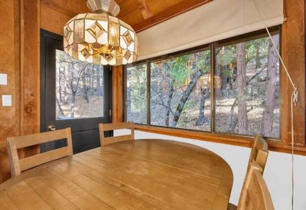 Une table à manger en bois, des chaises, un lustre et une vue sur les arbres à travers de grandes fenêtres.