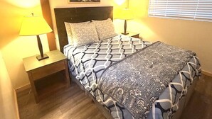 A bedroom with a bed, two bedside tables with lamps, a headboard, a painting on the wall, and a window with blinds.