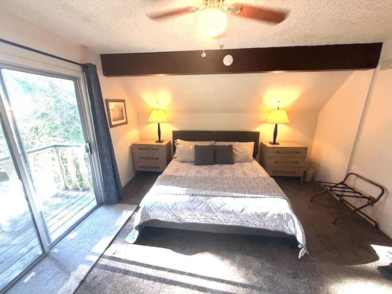 A bedroom with a bed, two bedside tables with lamps, a ceiling fan, a window, and a balcony.