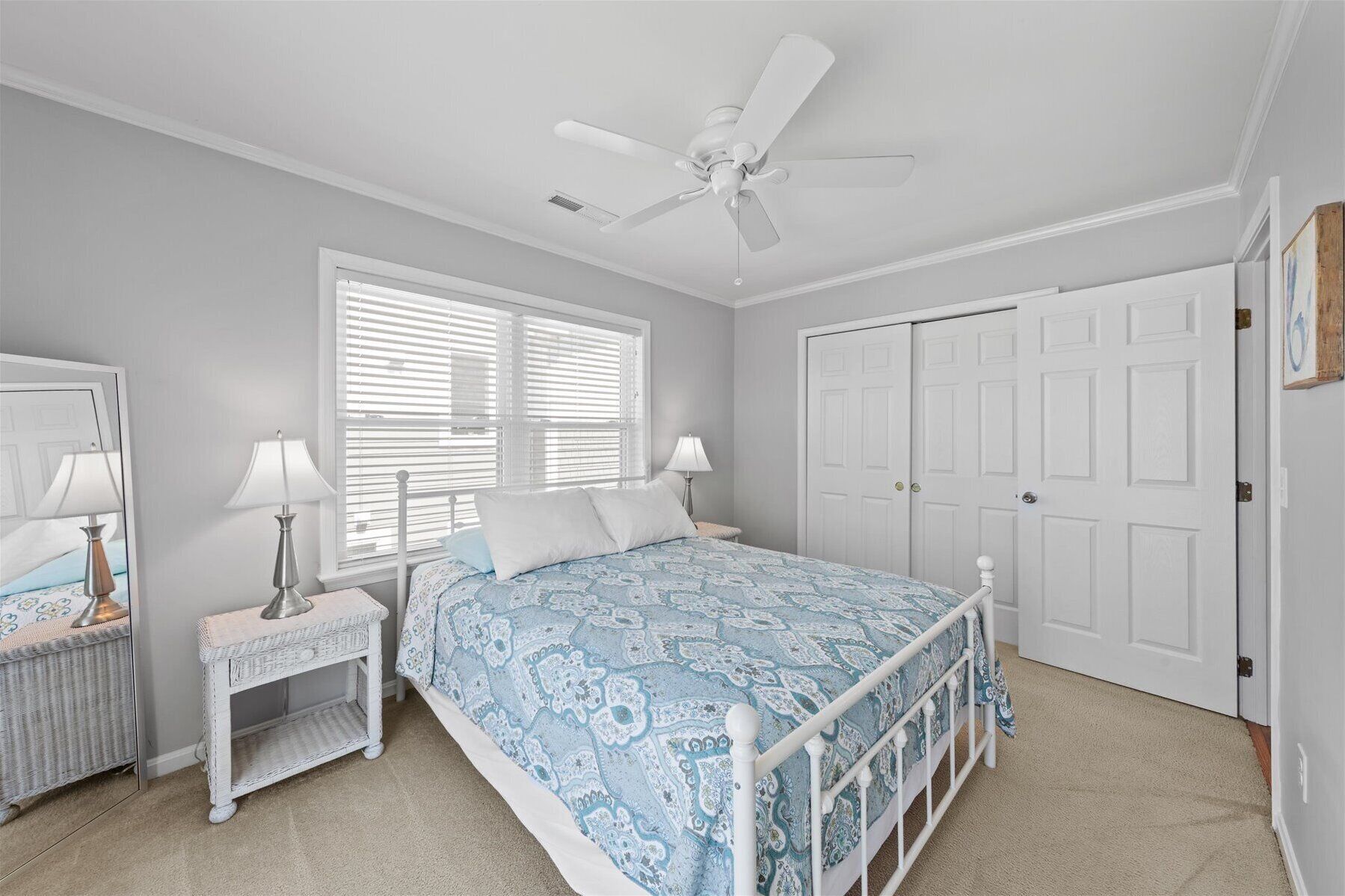 A bedroom with a bed, two bedside tables with lamps, a ceiling fan, a large window with blinds, and a wardrobe.