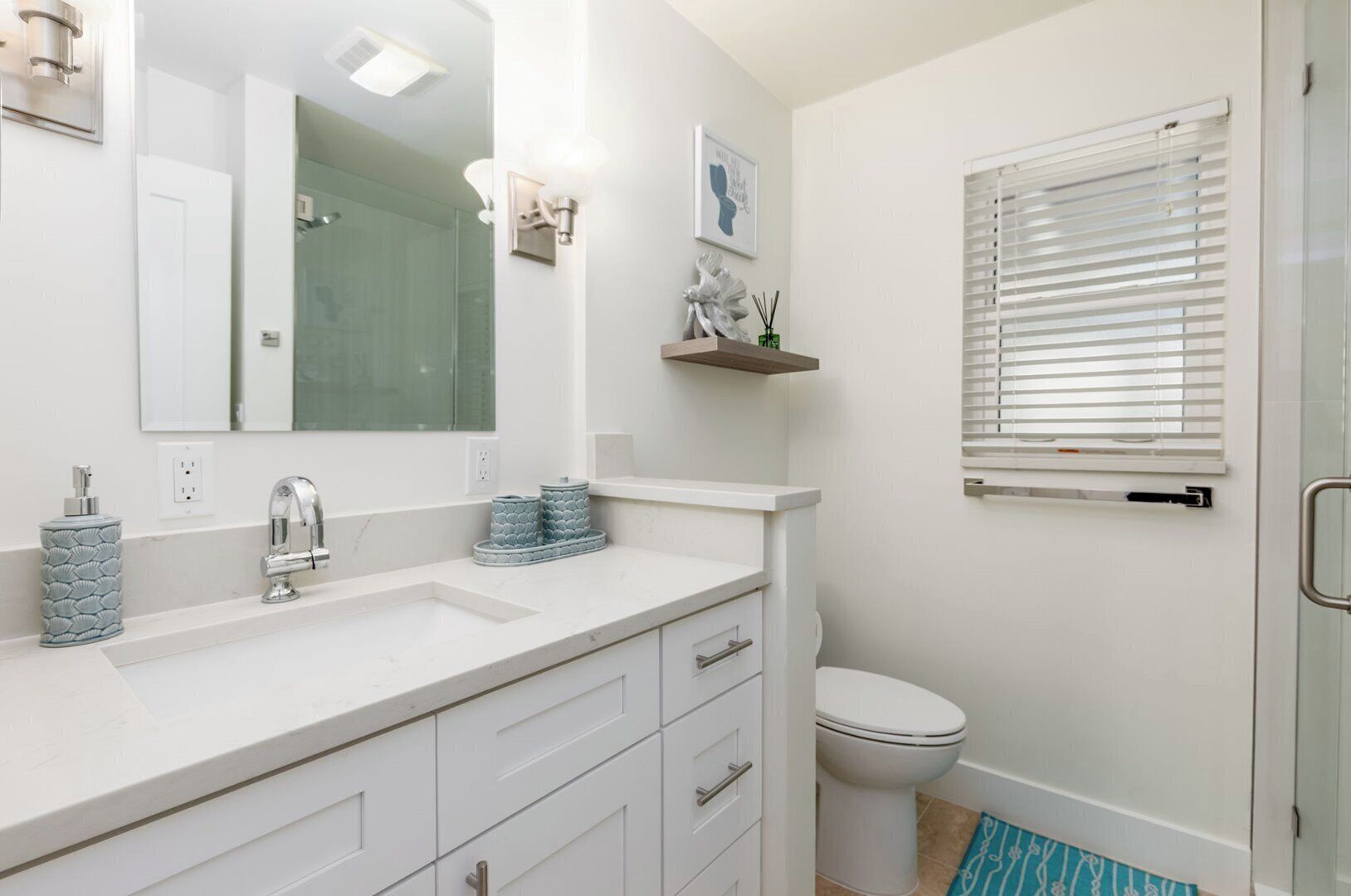 Ein modernes Badezimmer mit einem weißen Waschbecken, einem Spiegel und einer Toilette. Ein Fenster mit Jalousien und ein kleines Regal über der Toilette sind ebenfalls vorhanden.