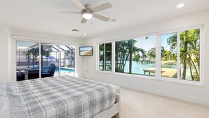 A bedroom with a bed, a ceiling fan, a TV, and large windows with a view of a pool and palm trees. - Water's Edge-Intracoastal Gem with Pool and Spa (Saint Petersburg)