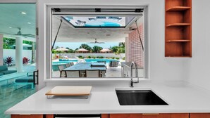 A modern kitchen with a large window overlooking a pool and outdoor seating area. - Casa Floridita by Duncan Real Estate (Anna Maria)