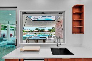 A modern kitchen with a large window overlooking a pool and outdoor seating area. - Casa Floradita by Duncan Real Estate (Anna Maria)