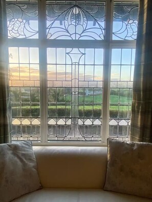 Una habitación con un gran ventanal con vidrieras intrincadas y celosías metálicas, un sofá blanco con dos cojines, y vistas a un paisaje verde.
