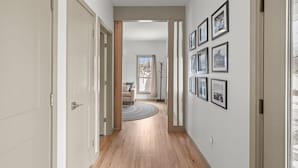 Un pasillo con suelo de madera, paredes blancas y cuadros enmarcados. Hay una puerta a la izquierda y una ventana al fondo.