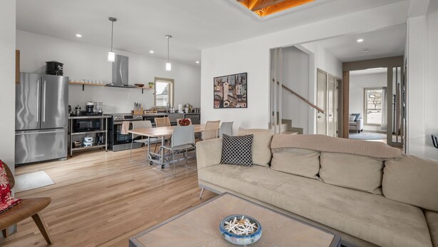 Una cocina moderna con electrodomésticos de acero inoxidable, un comedor con mesa y sillas blancas, y una sala de estar con sofá color beige y una mesa de centro.