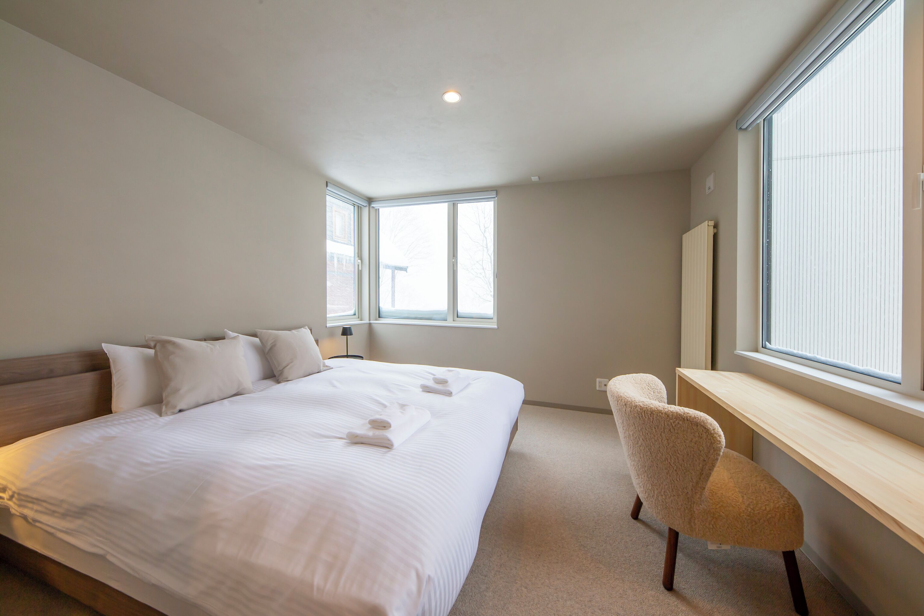 Un dormitorio con una cama grande, un escritorio y una silla. Dos ventanas permiten la entrada de luz natural.