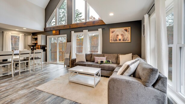 A spacious living area with a high ceiling, large windows, a sofa, a coffee table, and a dining area with chairs and a table. - White Mtns Holiday Home! Cabin 1 Mi to Skiing (Campton)