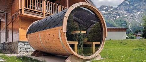Una estructura con forma de barril de madera y un ventanal de vidrio, ubicada en una zona cubierta de pasto con montañas al fondo.