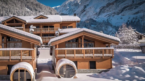 Ein Chalet-Stil Gebäude mit Holzbalkonen und großen Fenstern, umgeben von schneebedeckten Bergen.
