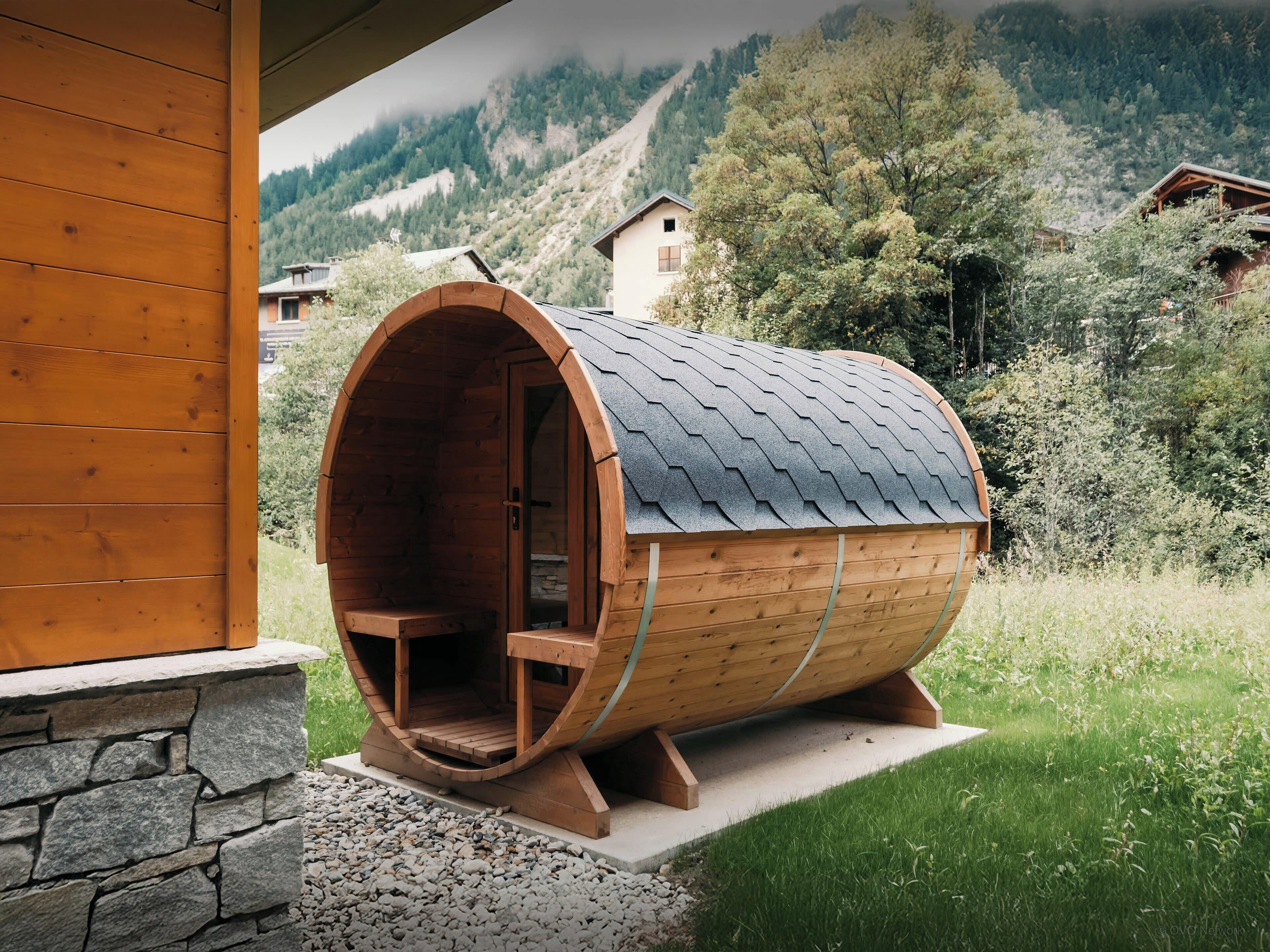 Une structure en bois, en forme de tonneau, avec un banc à l’intérieur, posée sur une base en pierre, entourée de verdure et avec une chaîne de montagnes en arrière-plan.