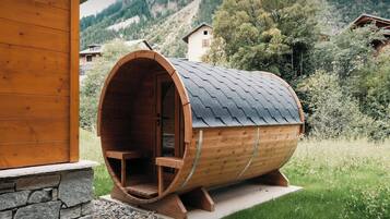 Une structure en bois, en forme de tonneau, avec un banc à l’intérieur, posée sur une base en pierre, entourée de verdure et avec une chaîne de montagnes en arrière-plan.