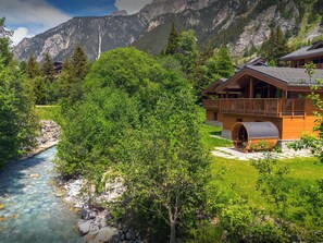 Un refugio de montaña con un río, vegetación exuberante y vistas a las montañas.