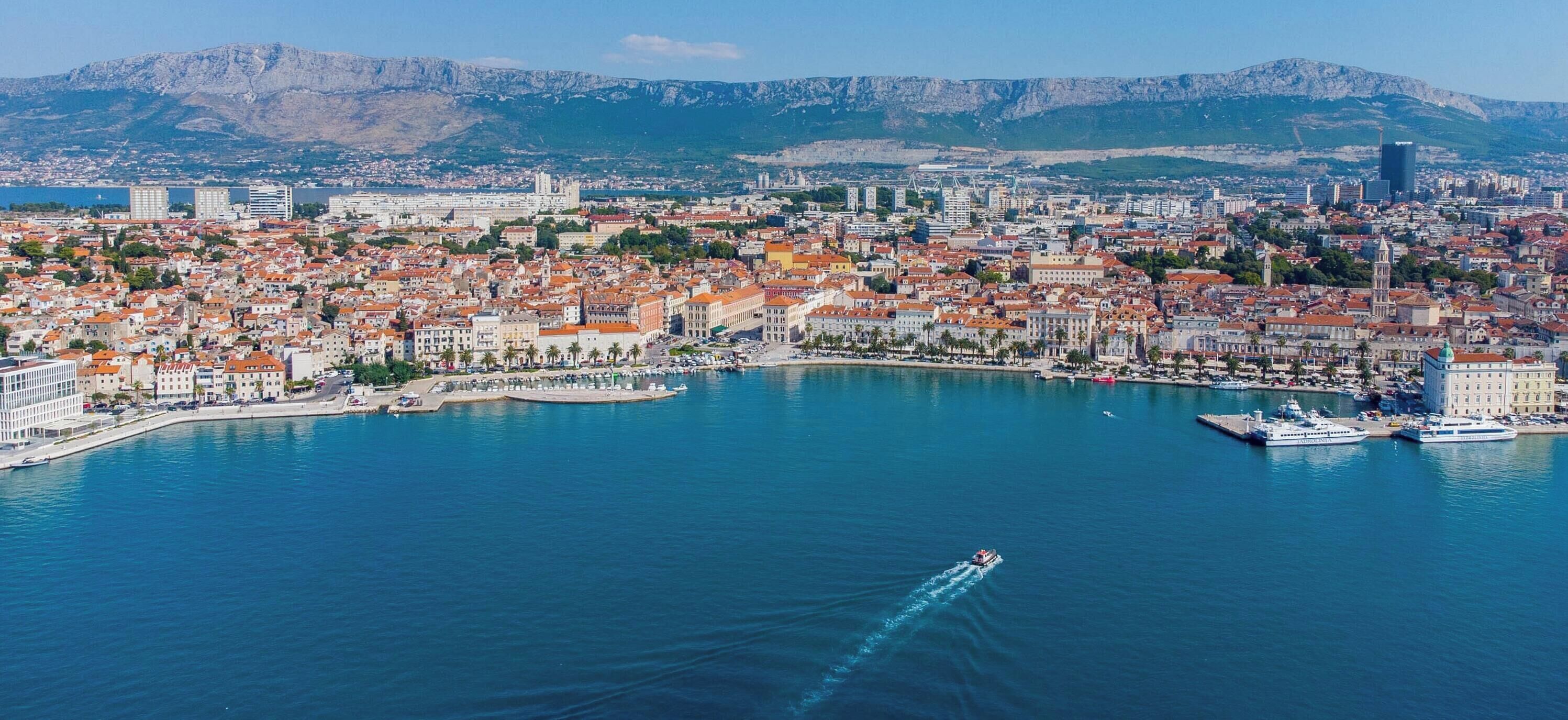Un paisaje costero con un puerto deportivo, edificios y montañas al fondo.