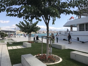 Eine Uferpromenade mit einem großen Baum, Sitzgelegenheiten und einem weißen Boot, das in der Nähe liegt.