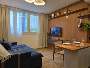 Ein modernes Zimmer mit einem Bett, einem Esstisch mit Stühlen, einem an der Wand montierten Fernseher und einem Fenster mit Vorhängen.