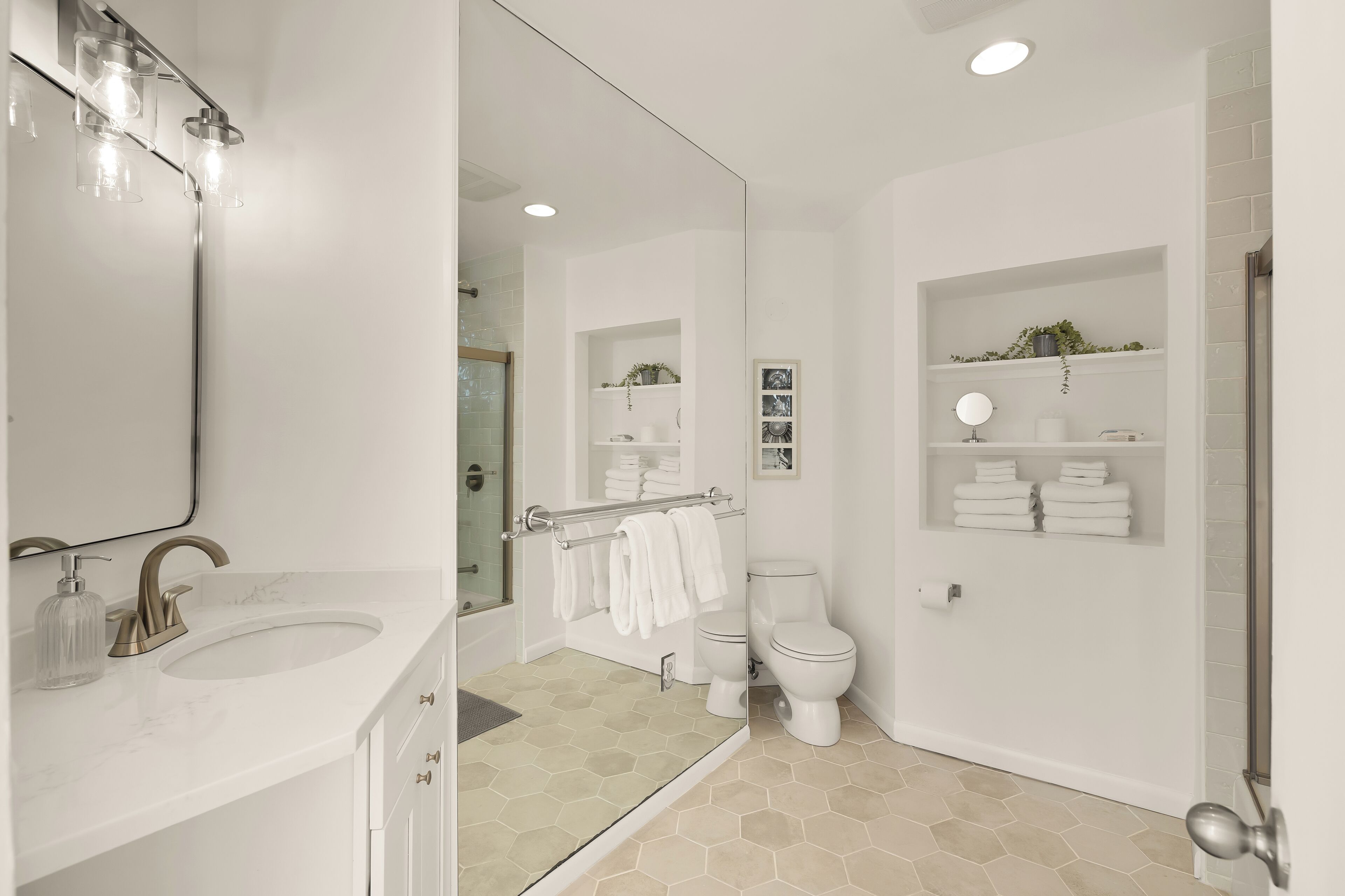 Une salle de bain moderne avec un grand miroir, un lavabo doté d’un robinet doré, un distributeur de savon en verre, une cabine de douche en verre, des toilettes et une étagère avec des serviettes et des articles de toilette.
