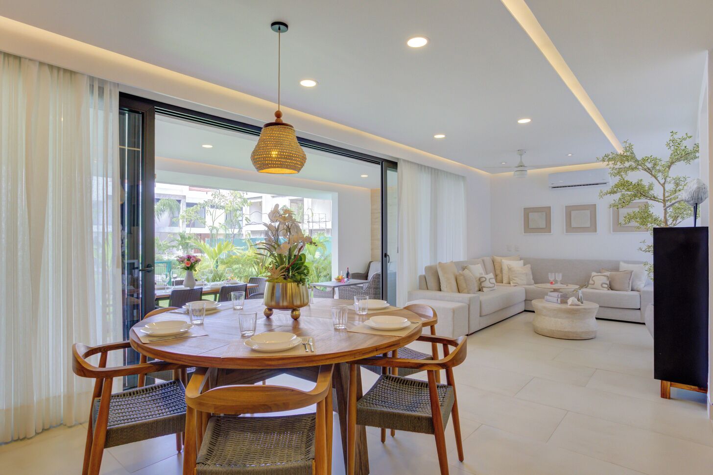 A modern living room with a dining area, featuring a round wooden table, wicker chairs, a hanging light fixture, a sofa, and a large window with a view of greenery.