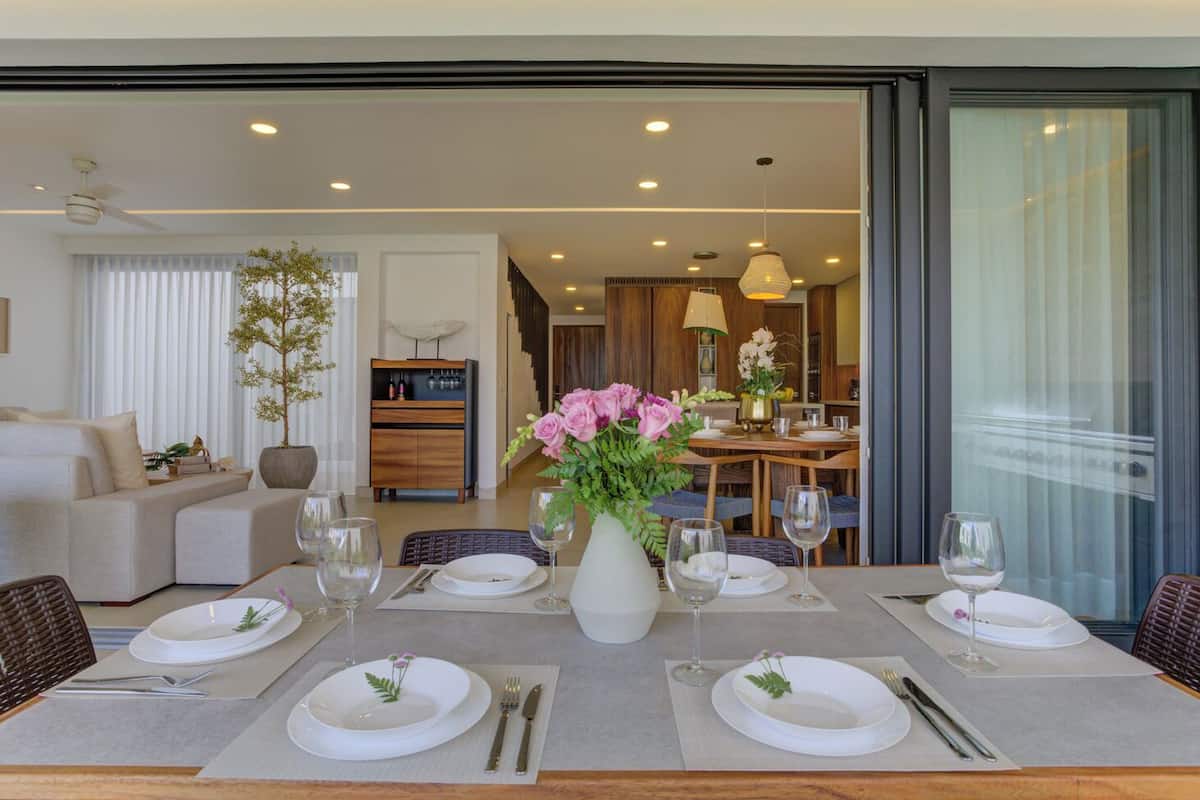 Un coin repas avec une table dressée pour quatre personnes, un vase de fleurs et, en arrière-plan, une vue sur le salon et la cuisine.