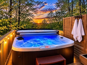 Un jacuzzi con cubierta de madera, rodeado de árboles, y un atardecer al fondo.
