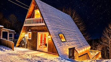 Een besneeuwde, driehoekige blokhut met een balkon en een verlicht interieur.