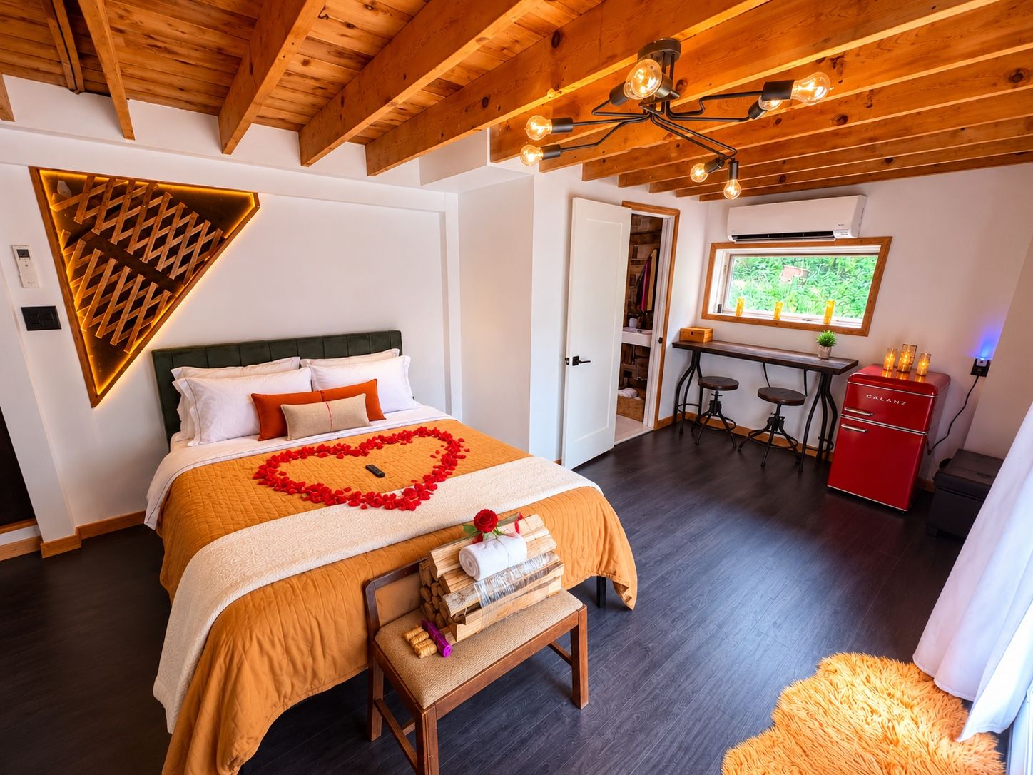 A hotel room with a bed, a desk, a red appliance, and a window with a view of greenery.