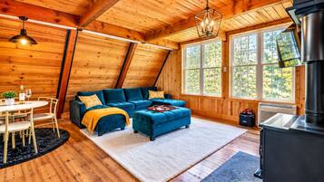 A cozy cabin interior with a wooden ceiling, a blue sofa set, a dining table with chairs, and a fireplace.