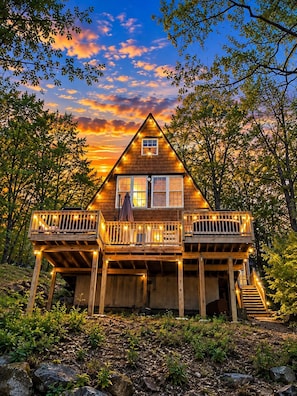 Una cabaña de madera con balcón, rodeada de árboles y una puesta de sol al fondo.