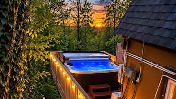 A hot tub on a wooden deck with string lights, surrounded by trees and a sunset in the background.