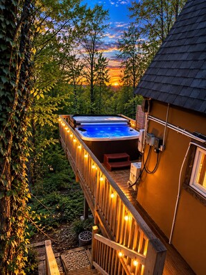 A hot tub on a wooden deck with string lights, surrounded by trees and a sunset in the background.