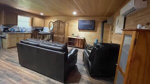 Un vaste espace de vie comprenant un grand canapé en cuir noir, des meubles en bois, un téléviseur fixé au mur et un coin cuisine avec des meubles en bois et un plan de travail.