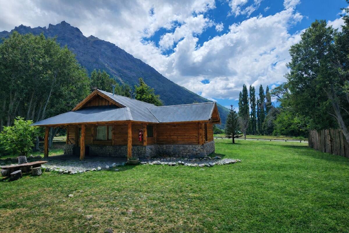 A wooden cabin with a stone foundation, surrounded by a grassy area and trees, with mountains in the background.
