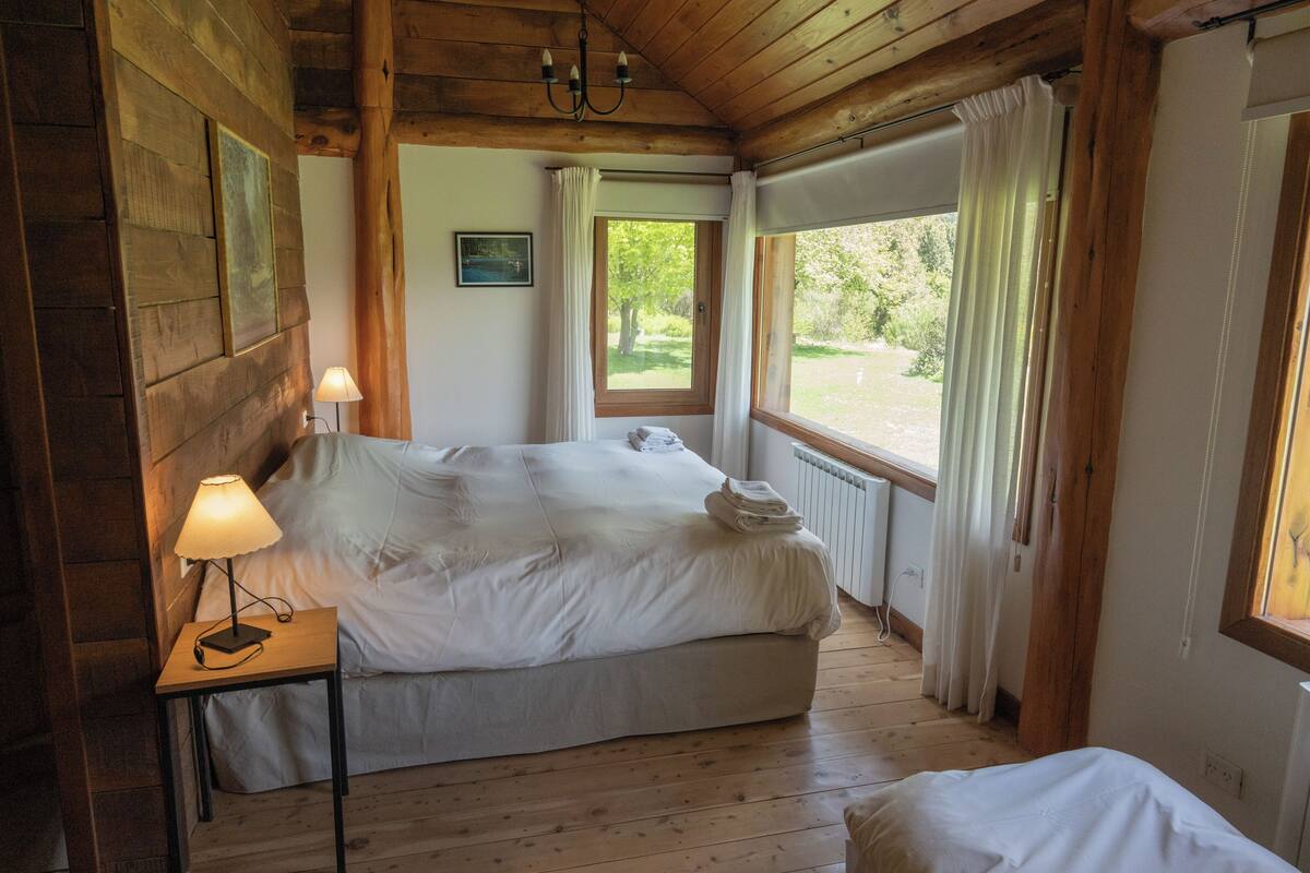 A wooden cabin bedroom with a bed, a nightstand with a lamp, a window with curtains, and a view of greenery outside.